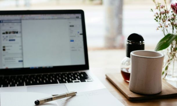 Laptop open with pen and notepad in front. Cup of coffee to the side and a vase of flowers.