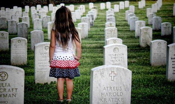 Young girl wearing a white, pink and blue dress standing in a cemetery with her back to camera