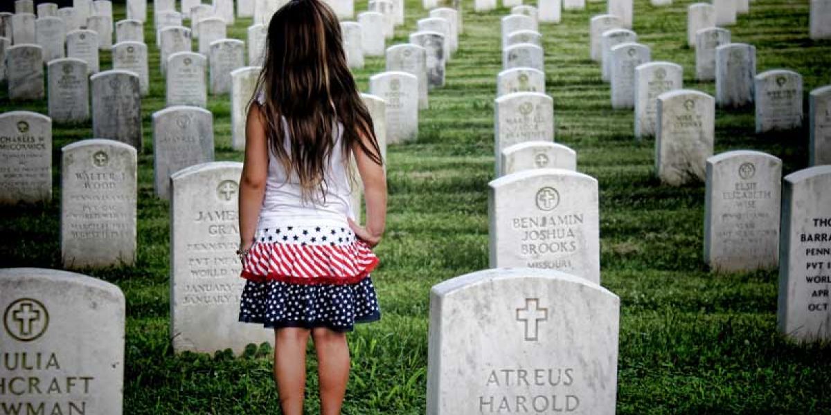 Young girl wearing a white, pink and blue dress standing in a cemetery with her back to camera